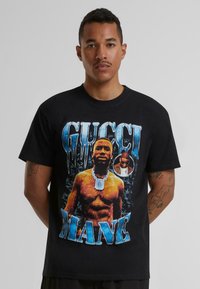 Black cotton t-shirt featuring a graphic print of a muscular figure with a chain, surrounded by icy blue lettering spelling "GUCCI MANE."