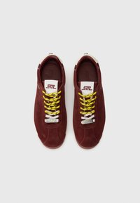 Un paio di sneaker in suede bordeaux con lacci bianchi e gialli, etichetta bianca sulla linguetta e punte dei lacci in metallo "Steve Madden", viste dall'alto.