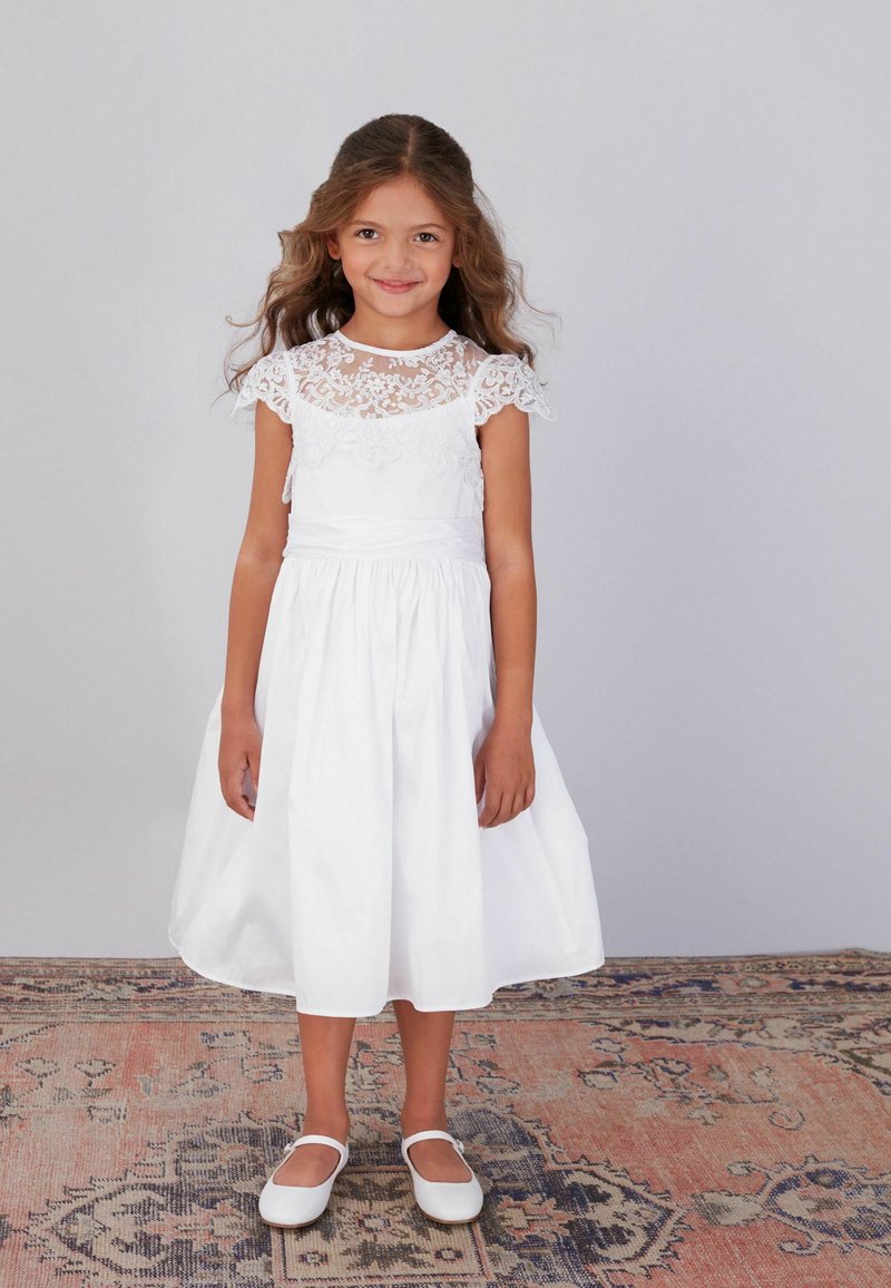 Niña joven con cabello largo y rizado que lleva un vestido blanco de encaje y zapatos blancos, de pie sobre una alfombra con estampado, frente a un fondo gris.