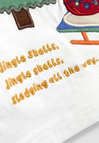 Jingle bells -lause broderad med orange tråd på vit tyg, nära en färgglad broderad gran och en figur som åker släde.
