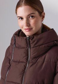 Pruun polsterdatud jakk kõrge kraega, millel on kaks tõmblukku ja tikandid. Kangas näib sile ja soe.