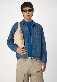 Chaqueta de mezclilla azul con un patrón intrincado, camiseta azul debajo, bolso de bandolera beige, pantalones cargo marrón claro y gafas de sol negras.