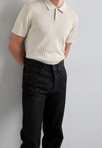 Vägsd beige-virkad polo med korta ärmar och krage, i kombination med mörka denimjeans. Skjortan har ett strukturerat räfflat mönster.