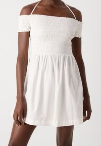 Bílé mini šaty s odhalenými rameny, smockovaným tělem, krátkými rukávy a halter krčním páskem, které nosí osoba s tmavou pletí.