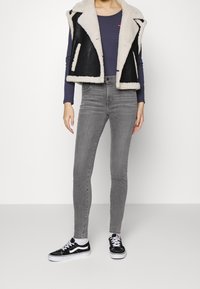 Svart och krämfärgad shearlingväst över en marinblå tröja, i kombination med grå tajta jeans och svarta sneakers med vita sulor och snörningsdetaljer.