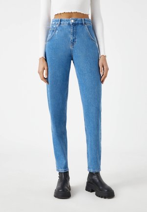 Donna che indossa jeans a vita alta azzurri chiaro con tasche frontali, abbinati a stivali neri robusti e una maglia bianca a maniche lunghe corta.