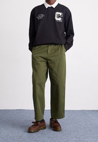 Schwarzes Polo mit langen Ärmeln, weißem Kragen und Brustlogo, kombiniert mit olivgrünen Hosen und braunen Schuhen mit Schnürdetails.