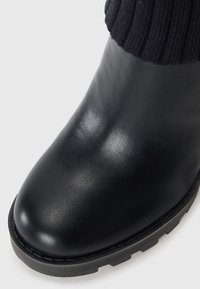 Black leather ankle boot with a rounded toe and a chunky rubber sole. Features a ribbed black knit sock-like upper for added warmth.