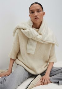 Donna che indossa un maglione di maglia color crema con una sciarpa abbinata drappeggiata sulle spalle, pantaloni grigi e ballerine beige, seduta su uno sfondo neutro.