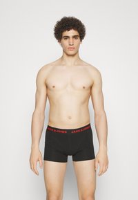 Schwarze Boxershorts mit rotem "JACK & JONES"-Bund, eng anliegendes Design, elastisches Material, getragen von einem Modell mit kurzen, lockigen Haaren.