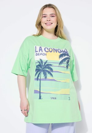 Camiseta oversized de color verde claro con un estampado gráfico de palmeras y una puesta de sol, con el texto "LA CONCHA BEACH" y "ESPAÑA". Mangas cortas.