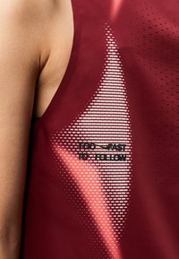 Κοντινή λήψη μπορντό αθλητικής φανέλας με λευκό και ροζ γραφικό σχέδιο και το κείμενο "TOO FAST TO FOLLOW" κοντά στη μασχάλη.