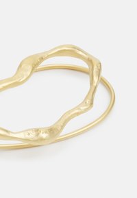 Goldfarbener Metallarmreif mit wavy, organischer Form, der ein strukturiertes, skulpturales Design und ein glattes, rundes Band aufweist.