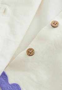 Gros plan sur un tissu crème avec deux boutons en bois et une petite zone de détails tricotés violets.