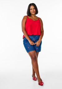 Czerwona bluzka bez rękawów z falbaniastym dekoltem, w parze z niebieskimi szortami dżinsowymi ozdobionymi haftem kwiatowym oraz czerwonymi balerinkami z ozdobami.