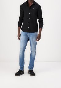 Czarna koszula zapinana na guziki z długimi rękawami, w połączeniu z jasnoniebieskimi dżinsami i czarnymi tenisówkami. Na koszuli znajduje się małe logo na piersi.