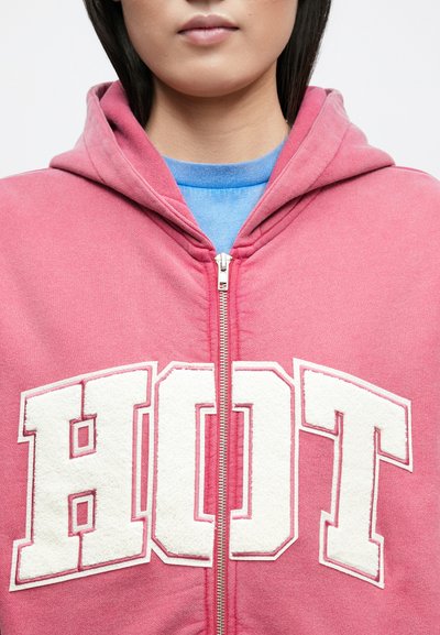 Rózsaszín cipzáras kapucnis pulóver fehér, bolyhos "HOT" felirattal elöl, állítható kapucnival és egy kék inggel alatta.