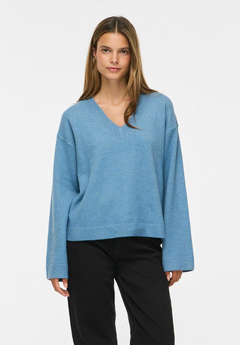Femme portant un pull ample bleu clair à col en V et un pantalon noir, debout sur un fond blanc uni.