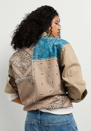 Vrouw met krullend haar die een beige en blauwe patchworkjas met bandana-patronen draagt, gecombineerd met een blauwe spijkerbroek, zijwaarts gezien.