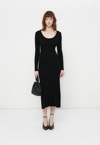 Black ribbed long-sleeve dress with a scoop neckline, straight silhouette, and mid-calf length. Paired with black heeled shoes and a small handbag.