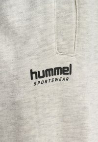Ljusgrå sportklädes tyg med en strukturerad yta, som har en svart "hummel"-logotyp och texten "SPORTSWEAR" nära en sidoficka.