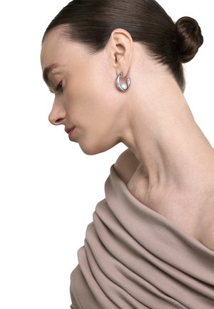 Moteris su tamsiais plaukais surištais kuodu, dėvi smėlio spalvos palaidinę be petnešėlių ir sidabrinį lankinį auskarą su perlu, profilio pozoje ant balto fono.