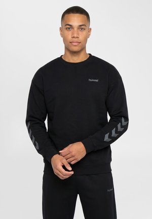 Ung mand iført en sort hummel sweatshirt med grå chevronmønstre på ærmerne og matchende sorte bukser på en hvid baggrund.