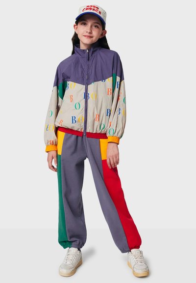 Farverig vindjakke med lilla top, beige midtersektion med multikolorede bogstaver, parret med grå sweatpants med røde, gule og grønne accenter.