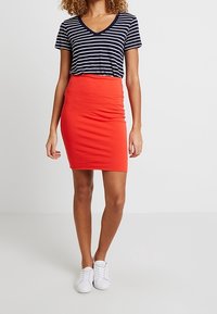 T-shirt à manches courtes à rayures bleu marine et blanches avec col en V, associé à une jupe crayon rouge ajustée. Mannequin portant des baskets blanches, debout sur un fond neutre.