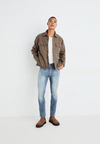 Chaqueta marrón de botones con dos bolsillos frontales, camiseta blanca, jeans de mezclilla azul claro y botas de ante marrones hasta el tobillo. Fondo liso.