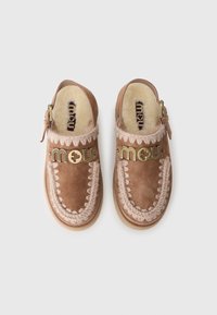Zoccoli in suede marrone con foderatura in pelliccia beige, cuciture decorative e un accento in metallo dorato con logo. Chiusura con fibbia laterale per una vestibilità regolabile.