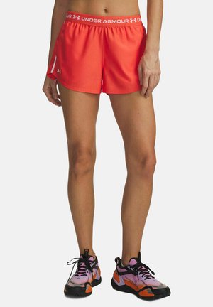 Vrouw die feloranje sportshorts van Under Armour draagt en meerkleurige hardloopschoenen in paars, oranje en zwart, staande tegen een witte achtergrond.