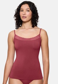 Kobieta z kręconymi włosami, ubrana w czerwony top bez rękawów z koronkowym wykończeniem i pasującą bielizną, stojąca na jednolitym tle.