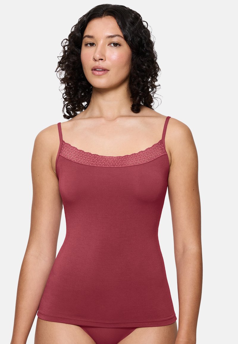 Kobieta z kręconymi włosami, ubrana w czerwony top bez rękawów z koronkowym wykończeniem i pasującą bielizną, stojąca na jednolitym tle.