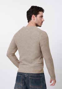 Pull beige en maille avec des accents côtelés sur les manches, une finition texturée et un col rond. Porté avec un pantalon en denim bleu.