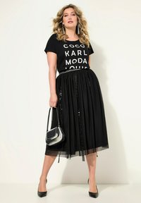 La maglietta grafica nera presenta un audace testo bianco. È accompagnata da una gonna in tulle nera con dettagli in paillettes, una borsa argentata e tacchi neri.
