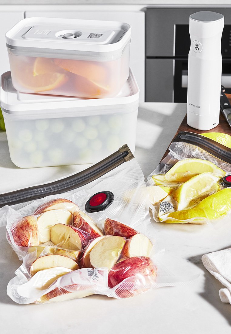 Vakuumverpackte Beutel mit Apfel- und Zitronenscheiben, transparent mit roten und schwarzen Ventilen; zwei klare Behälter mit weißen Deckeln, gestapelt.