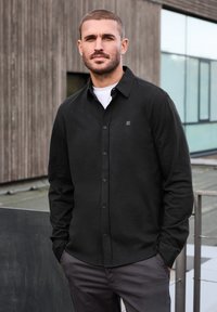 Svart långärmad skjorta med knappar och en diskret logotyp, tillverkad av ett mjukt material, i kombination med grå byxor i en avslappnad utomhusmiljö.