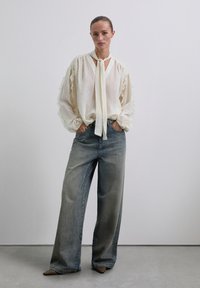 Blusa crema con mangas acampanadas, cuello atado, combinada con jeans de pierna ancha azul desgastados. Textura suave, diseño casual, colores neutros.