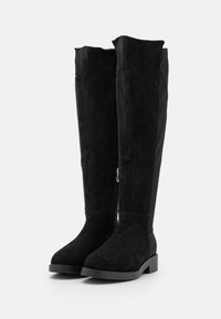 Botas negras de ante por encima de la rodilla, con suela plana y cremallera lateral para abrochar. Acabado suave con un aspecto mate.