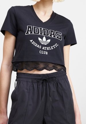 Kobieta nosząca czarną krótką koszulkę Adidas z dekoltem w serek z białym logo i napisem, nałożoną na czarną bluzkę z koronkowym wykończeniem, zestawioną z czarnymi spodniami z troczkiem.
