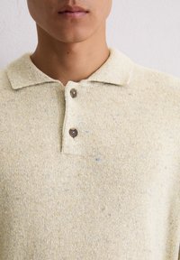 Beige gebreide polo met een textuurpatroon, voorzien van een kraag, twee bruine knopen en subtiele blauwe stippen door de stof.