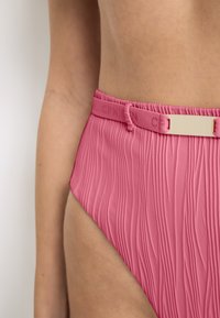 Záběr zblízka na osobu nosící růžové texturované plavky s vysokým pasem a malým nastavitelným páskem v pase.