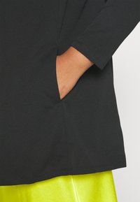 La main d'une personne dans la poche d'un vêtement noir à manches longues, avec une jupe ou un pantalon jaune néon vif visible en dessous.
