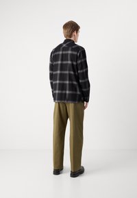 Černá a šedá kachlová flanelová košile s volným střihem, kombinovaná s olivově zelenými kalhotami. Outfit doplňují černé boty a neutrální pozadí.