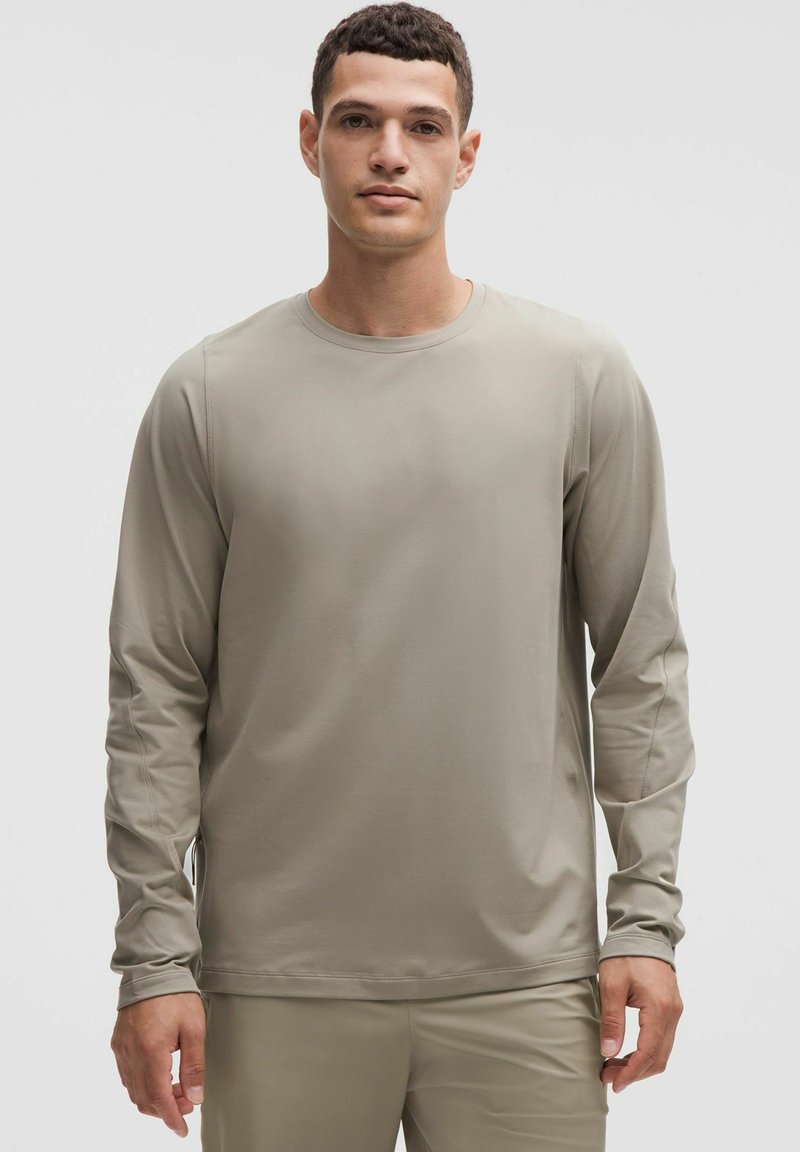 Cămășă lungă cu mâneci, gri deschis, textură netedă, decolteu rotund, croi drept, cu detalii de cusătură subtile pe mâneci.