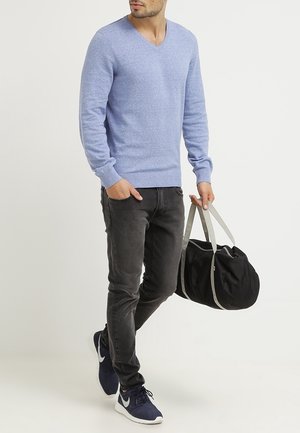 Hellblauer Pullover mit V-Ausschnitt, dunkelgraue Jeans und marineblaue Sneaker. Hält eine schwarze Reisetasche mit grauen Trägern. Schlichtes, lässiges Design.