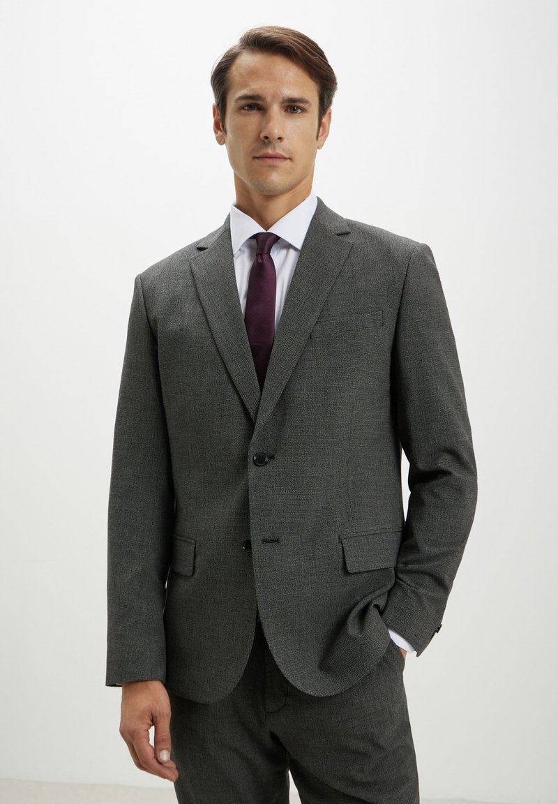 Costume gris foncé présentant un motif texturé discret, avec des revers en notch, deux boutons et deux poches avant. Porté avec une chemise blanche et une cravate bordeaux.