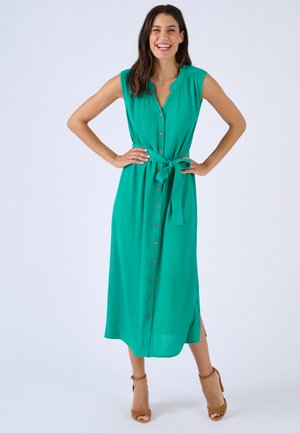 Donna sorridente in abito verde senza maniche con bottoni e cintura in vita, con sandali marroni con tacco, in piedi con le mani sui fianchi.