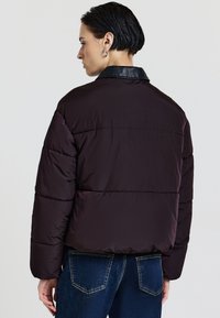 Dunkelviolette Daunenjacke mit gestepptem Design und schwarzem Lederkragen. Sie hat lange Ärmel und wird mit blauen Jeans kombiniert.